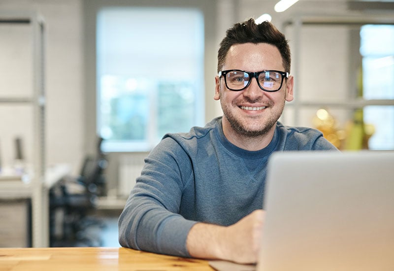 An employee wearing glasses, sitting in front of a laptop and smiling because they received positive feedback via email.