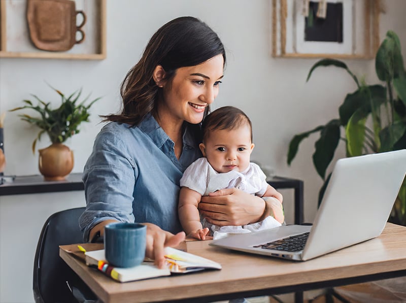 A working mother holds her baby while seated at her desk.